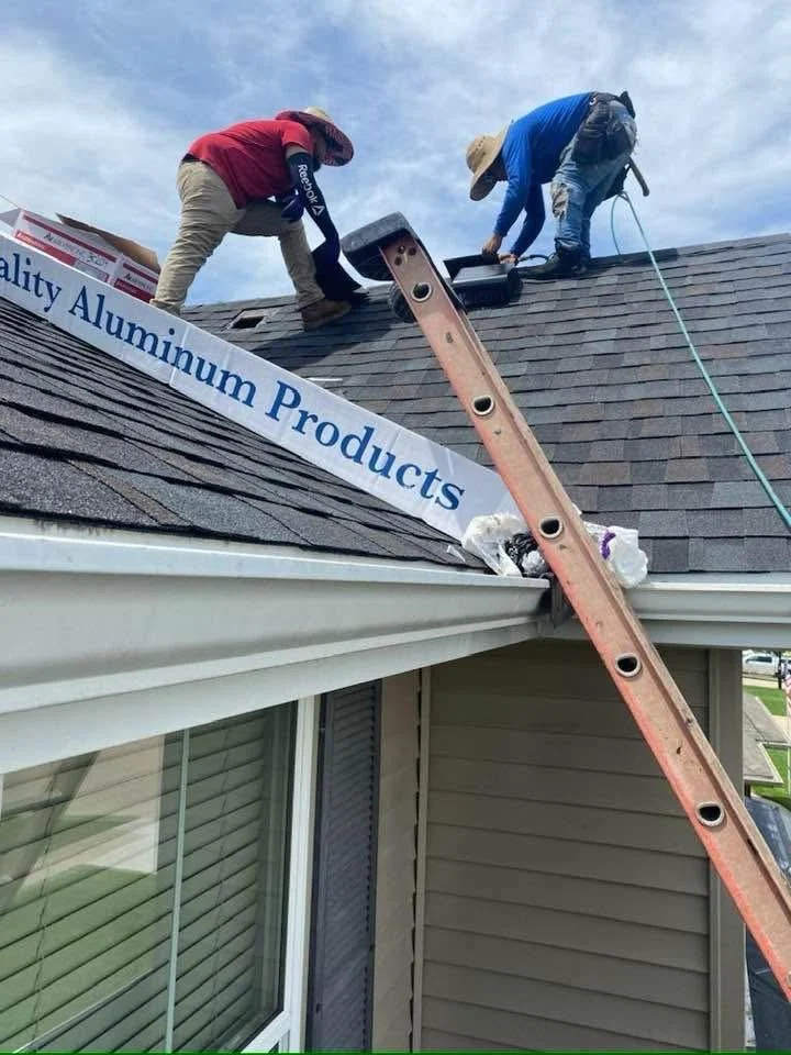 Two roofers working above a ladder at the edge of a shingle roof